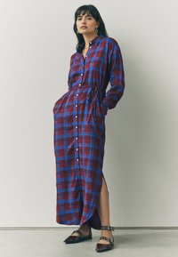 Lang ternet kjole i rød, blå og hvid. Har knaplukning foran, lange ærmer, bindebånd i taljen og slidser i siden. Båret med spidse flade sko.