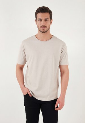Homme aux cheveux courts et foncés avec une barbe, portant un t-shirt beige clair uni et un pantalon noir, debout devant un fond blanc.