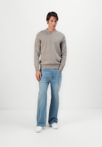 Suéter de malha cinza claro com decote redondo, combinado com jeans azuis abertos e tênis brancos, contra um fundo neutro.