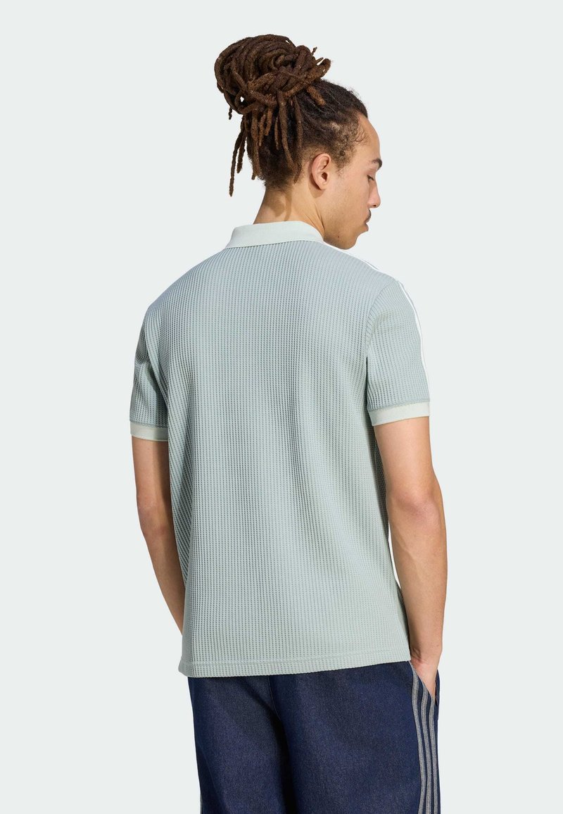 Mann mit Dreadlocks, der ein hellgraues, strukturiertes Kurzarm-Poloshirt und dunkelblaue Hose trägt, von hinten gesehen mit Händen in den Taschen.