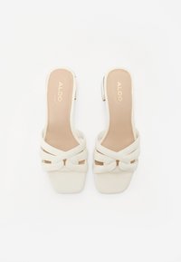 Valkoiset slingback-sandaalit neliön muotoisella varvastyöllä, ristikkäisvaljailla ja teksturoidulla pohjalla, jossa on hienovarainen logo sisäpohjassa.