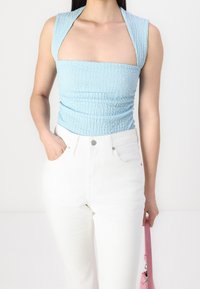 Woman wearing textured light blue sleeveless top and high-waisted white pants, holding a pink handbag in one hand, other hand in pocket.