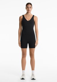 Black sleeveless bodysuit with a V-neck, fitted design, and short legs. Model wears white sneakers and ankle socks. Smooth texture.