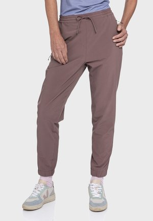Personne portant un pantalon de jogging mauve à taille élastique avec cordon et des baskets grises avec des lacets blancs, debout avec une main sur la hanche et l'autre sur la cuisse.