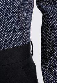 Námořnický pulovr s bílým cikcakovým vzorem, žebrovaným černým pasem a logem. Měkká textura a přiléhavý design s ležérním vzhledem.