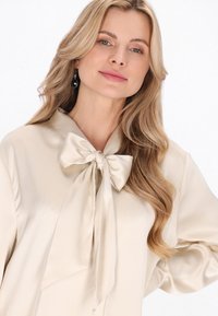 Beige satinbluse med stor sløjfe ved kraven, lange ærmer og glat tekstur, sat mod en lys baggrund.