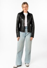 Zwarte leren jack met een ritssluiting aan de voorkant, een lichte kraag en een getailleerd ontwerp. Gepaard met lichtblauwe wijde spijkerbroeken en grijze sneakers.