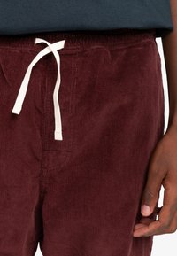 Bordeaux cordfløjl bukser med tekstureret stof, der har en hvid snøre og en detalje med baglomme. Overdelen er en navy t-shirt.