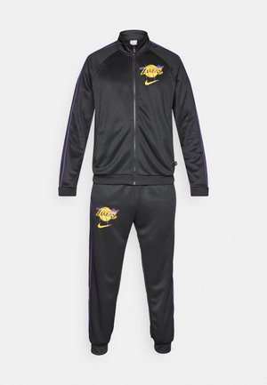 Tuta nera con accenti viola. Comprende una giacca con zip con logo dei Lakers e pantaloni abbinati, che mostrano un design elegante e sportivo.