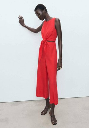 Une combinaison rouge avec des jambes larges et un nœud à la taille. Tissu lisse, design sans manches, avec un détail découpé. Des talons noirs complètent le look.