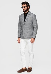 Homme portant un blazer gris, une chemise noire, un pantalon blanc, des mocassins marron et des lunettes de soleil noires, debout sur un fond blanc uni.