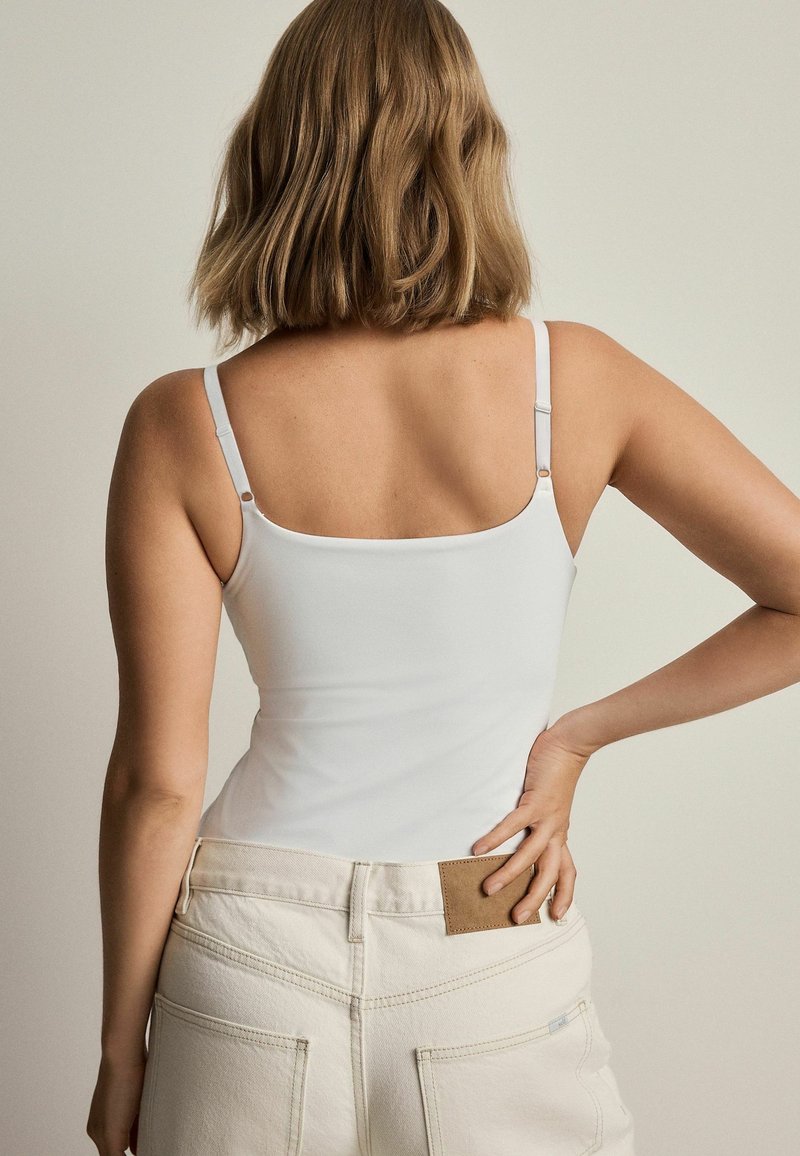 Femme aux cheveux mi-longs portant un débardeur blanc et un pantalon clair taille haute, main posée sur la hanche.
