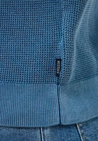 Blue textured knit sweater hem with a small black label reading "MUSTANG" above blue denim jeans.