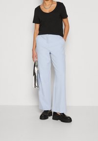 Osoba ubrana w jasnoniebieskie szerokie spodnie, czarną bluzkę z krótkim rękawem, czarne masywne loafersy, srebrny naszyjnik łańcuszkowy, trzymająca metaliczną srebrną torbę.