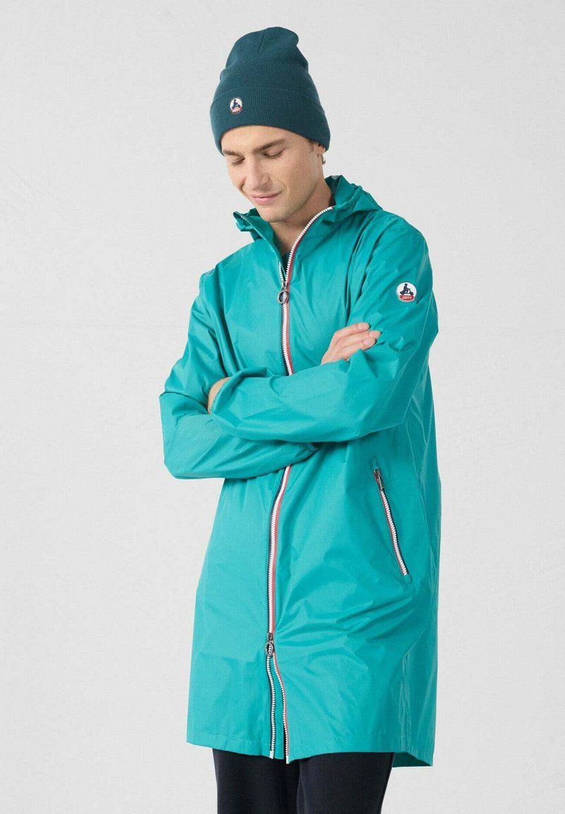 Giacca impermeabile turchese con cerniera frontale, finiture rosse a contrasto, tasche laterali e toppa con logo sulla manica. Abbinata a un berretto scuro.
