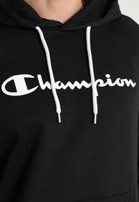 Felpa nera con cappuccio e lacci bianchi, caratterizzata da un grande logo bianco "Champion" sul petto in un font corsivo e una tasca frontale.