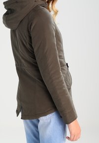 Oliwkowo-zielona kurtka z kapturem, zapinana na guziki z przodu, z kieszeniami po bokach i dopasowanym fasonem. Miękka tkanina o gładkiej fakturze, lekko taliowana.