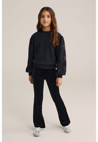 Črna majica s pufastimi rokavi in z zvezdnimi detajli; v kombinaciji z črnimi širokimi hlačami s belimi pikami. Sivo športno obutev dopolnjuje videz.