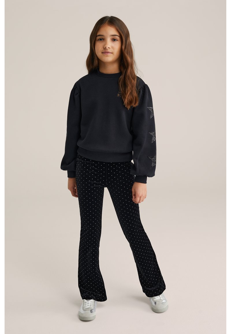 Črna majica s pufastimi rokavi in z zvezdnimi detajli; v kombinaciji z črnimi širokimi hlačami s belimi pikami. Sivo športno obutev dopolnjuje videz.
