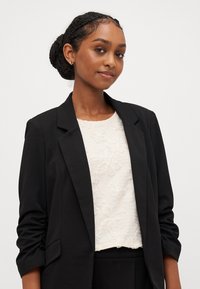 Μαύρο σακάκι κομμένο πάνω από λευκή υφασμάτινη μπλούζα με λουλουδάτα σχέδια. Το σακάκι έχει δομημένο σχέδιο και γυρισμένα μανίκια.