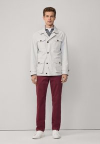 Veste gris clair avec quatre poches avant et boutons bruns, associée à un pantalon bordeaux et des baskets blanches, se tenant devant un fond neutre.