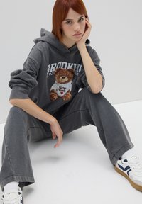 Felpa grigia in cotone con grafica di orso e testo "Brooklyn", abbinata a pantaloni jogger coordinati. Modello seduto, con sneakers bianche.