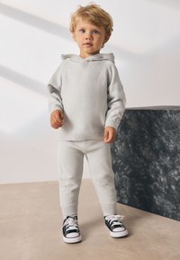 Sudadera con capucha gris claro y texturizada con pantalones a juego y puños acanalados. El niño lleva zapatillas negras y blancas, de pie sobre una superficie beige claro.