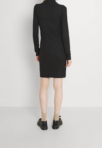 Svart ribbad långärmad klänning med hög halsringning, smal passform och nederkant som slutar ovanför knäet. Bärs med svarta skor och beige strumpor.