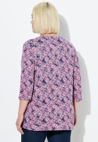 Donna con capelli biondi a caschetto che indossa un top a maniche lunghe con motivo floreale blu navy e rosa e jeans scuri, voltata di spalle contro un muro bianco.