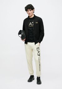 Čierna bunda na zips, zlaté tričko s logom, krémové tepláky s vertikálnou potlačou a čierne tenisky, držiac čiernu tašku.