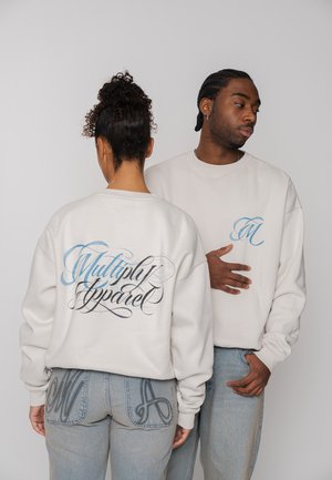 Frau und Mann tragen weiße Sweatshirts mit blauer und schwarzer Kalligraphie; die Frau steht mit dem Rücken zur Kamera und zeigt den Schriftzug "Multiply Apparel", während der Mann seitlich schaut.