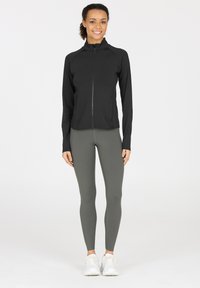 Zwarte ritsjack met een hoge kraag en lange mouwen, gecombineerd met hooggerokte, slim-fit grijze leggings. Witte sportschoenen.