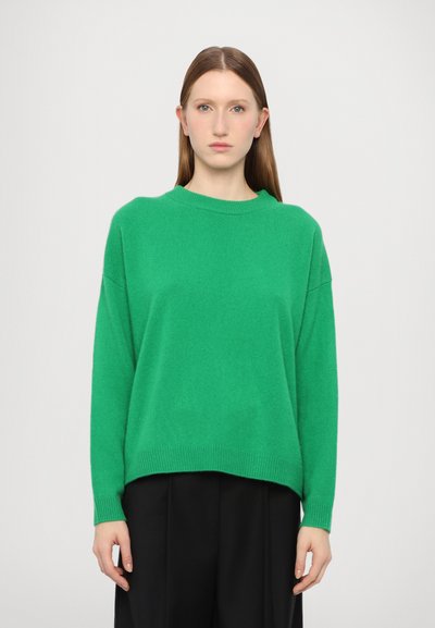 Suéter verde hecho de un suave material de punto, con cuello redondo, hombros caídos y dobladillo acanalado. Llevado con pantalones negros.