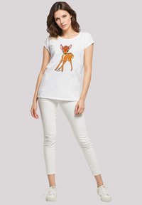 White t-shirt featuring an orange cartoon fawn design, short sleeves, and a relaxed fit, paired with white skinny jeans and white sneakers.
