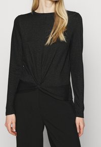 Svart långärmad topp i glittrig tyg med en tvinnad framsidesdesign och en slank silhuett. Matchas med svarta byxor.