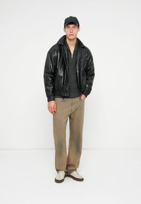 Svart skinnbomberjakke, grå zip-up genser, khaki vide bukser og hvite joggesko med brune detaljer. Modellen har på seg en svart caps.