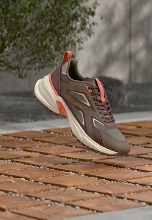 Bruin en koraalrode Calvin Klein sneaker zwevend boven een buitenpad van bakstenen en kiezelstenen met een wazige achtergrond van groen en een muur.