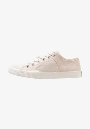 Beige lage canvas sneaker met witte rubberen zool, metalen oogjes en veters, met subtiele branding aan de hielzijde.