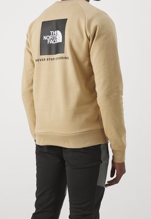 Osoba nosząca beżową bluzę z czarnym logo "The North Face" i napisem "Never Stop Exploring" na plecach oraz czarne spodnie z szarymi akcentami.