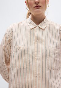 Ανοιχτό ροζ πουκάμισο με γιακά και κάθετες ρίγες, με δύο τσέπες μπροστά και κουμπιά κλεισίματος. Λείο ύφασμα με ελαφρώς γυαλιστερή υφή.