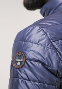 Giacca puffer blu navy con design trapuntato, dotata di una patch rotonda con logo sulla spalla. Tessuto lucido e texturizzato con colletto aderente.