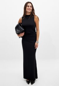 Abito nero senza maniche con colletto alto, arricciature in vita e silhouette aderente. Abbinato a una pochette nera e tacchi a punta.