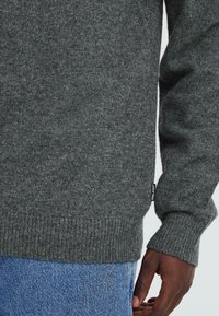 Grau gestrickter Pullover aus weichem Stoff, mit einem gerippten Saum und Bündchen, sowie einem kleinen Logotag an der Seite. Kombiniert mit hellblauen Jeans.
