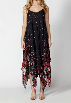 Kobieta ubrana w czarną sukienkę bez rękawów z białymi i czerwonymi kwiatowymi wzorami oraz asymetrycznym dołem, w połączeniu z sandałami na przezroczystych paskach.
