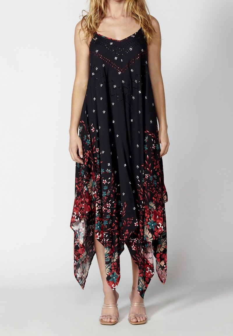 Femme portant une robe noire sans manches avec des motifs floraux blancs et rouges et un ourlet asymétrique, associée à des sandales à brides transparentes.