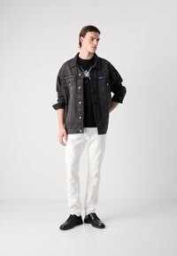 Zwarte denimjacket met knoopjes, gelaagd over een zwart T-shirt met een logo, gecombineerd met witte broek en zwarte sneakers.