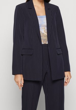 Námořnický pruhovaný blazer vyrobený z pevných textilií. Obsahuje dvě přední kapsy, výstřih do "v" a uvolněný střih. Nosený přes vzorovaný top.