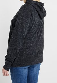 Sudadera gris oscuro con una tela texturizada, que presenta capucha, mangas largas y un corte relajado. Notables motas de color en toda la prenda.