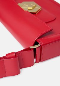 Borsa a mano in pelle rossa con una trama liscia, dettagli geometrici in oro e design piegato. Presenta una tracolla ampia con un dettaglio a fiocco.
