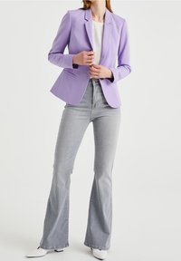 Žena oblečená v lila sakó přes bílé tričko s lehce šedými zvonovými džíny a bílými špičatými botami na pozadí prosté bílé stěny.